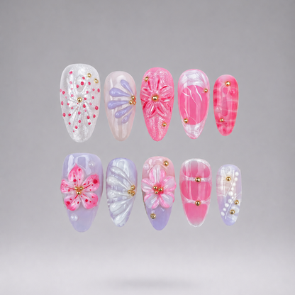 Ten false press-on nails arranged in two rows on a smooth gray gradient background. The nails feature various pink and pastel designs, including floral patterns, marble textures, glitter details, and small gold bead accents.