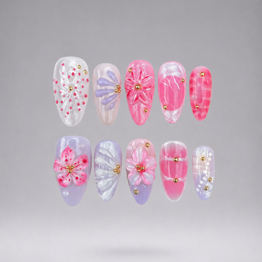 Ten false press-on nails arranged in two rows on a smooth gray gradient background. The nails feature various pink and pastel designs, including floral patterns, marble textures, glitter details, and small gold bead accents.