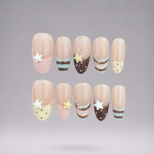 Ten false press-on nails arranged in two rows against a soft gray gradient background. The nails feature a nude beige base with pastel pink tips, decorative white stars, yellow stars, brown stripes, and light blue stripes.